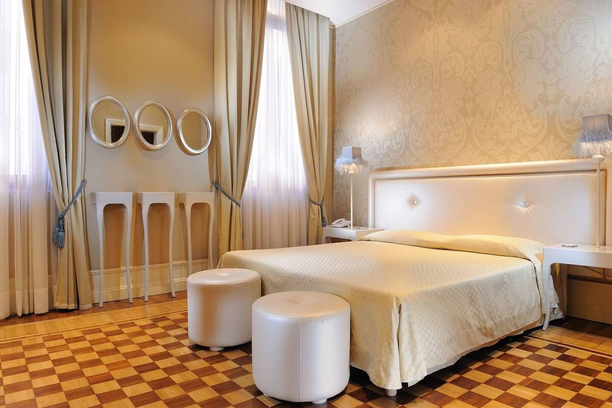 Quarto de hotel com chão xadrez, uma cama grande com cabeceira branca, duas mesas de cabeceira e três espelhos fixados na parede.