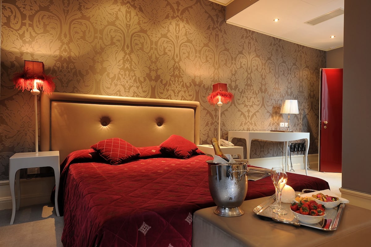 Quarto de hotel com uma cama grande, roupa de cama vermelha, uma mesa de cabeceira com uma bandeja de comida e uma garrafa de champanhe, e duas luminárias.