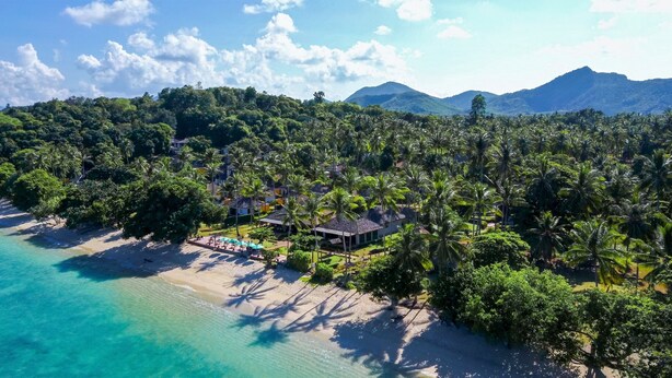 An aerial view of a tropical resort with a beachfront, dense palm tree coverage, and a mountain backdrop. - Elixir Koh Yao Yai (Ko Yao)