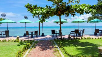 Un patio frente al mar con mesas y sillas bajo sombrillas verdes, con vistas al océano.