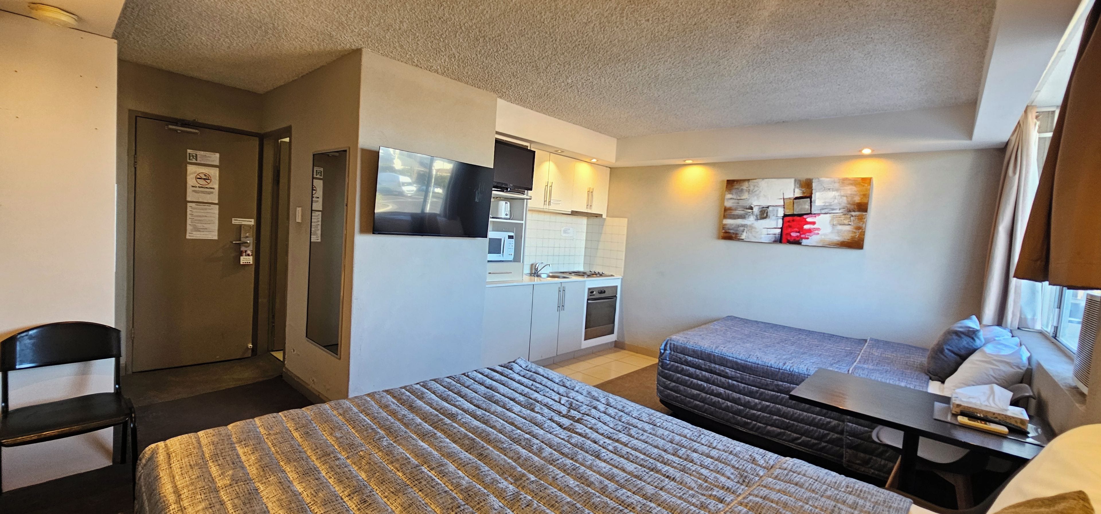 A compact hotel room with two beds, a kitchenette, a small table, and a window with a view.