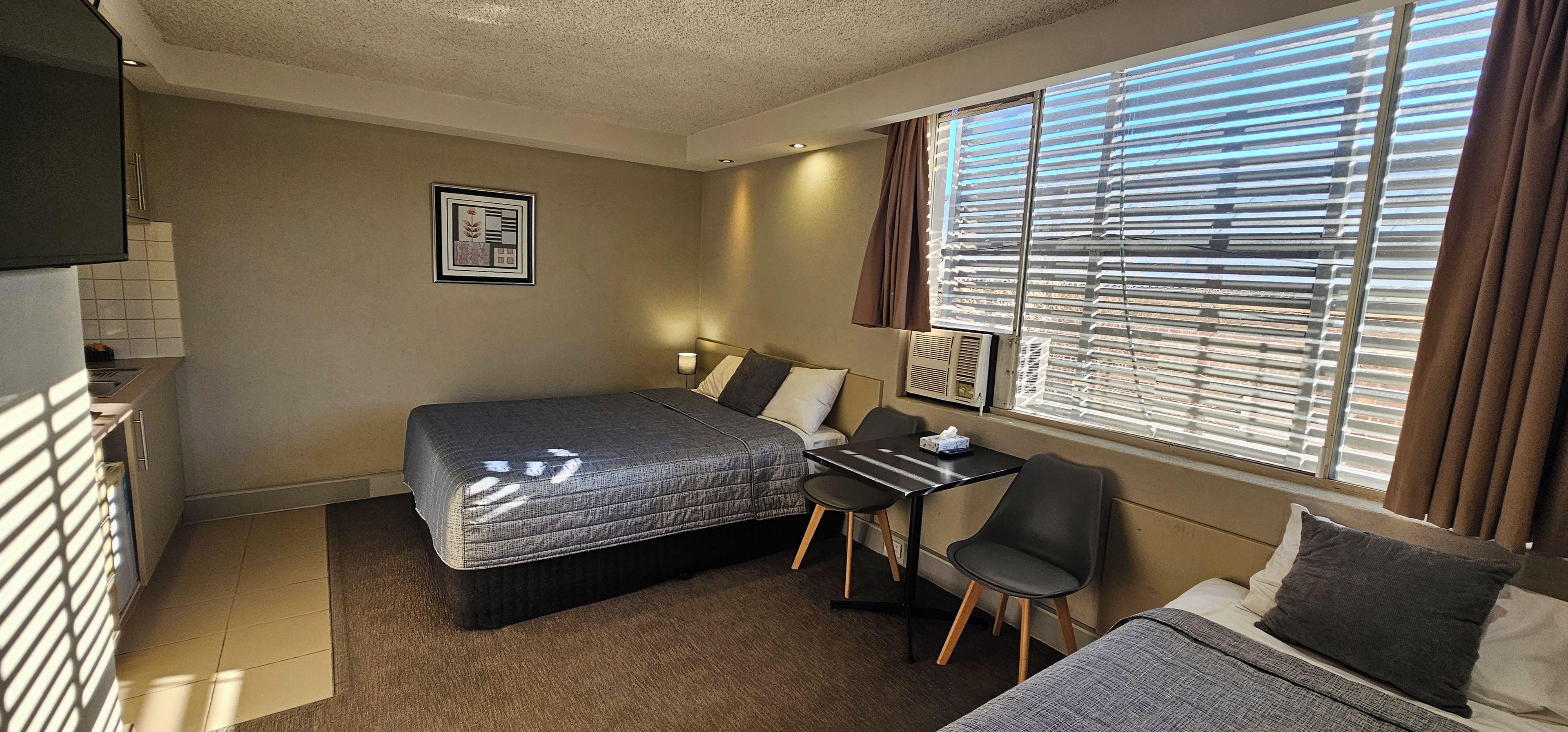 Ein kleines Hotelzimmer mit Bett, Schreibtisch, Stuhl und einem Fenster mit Jalousien.