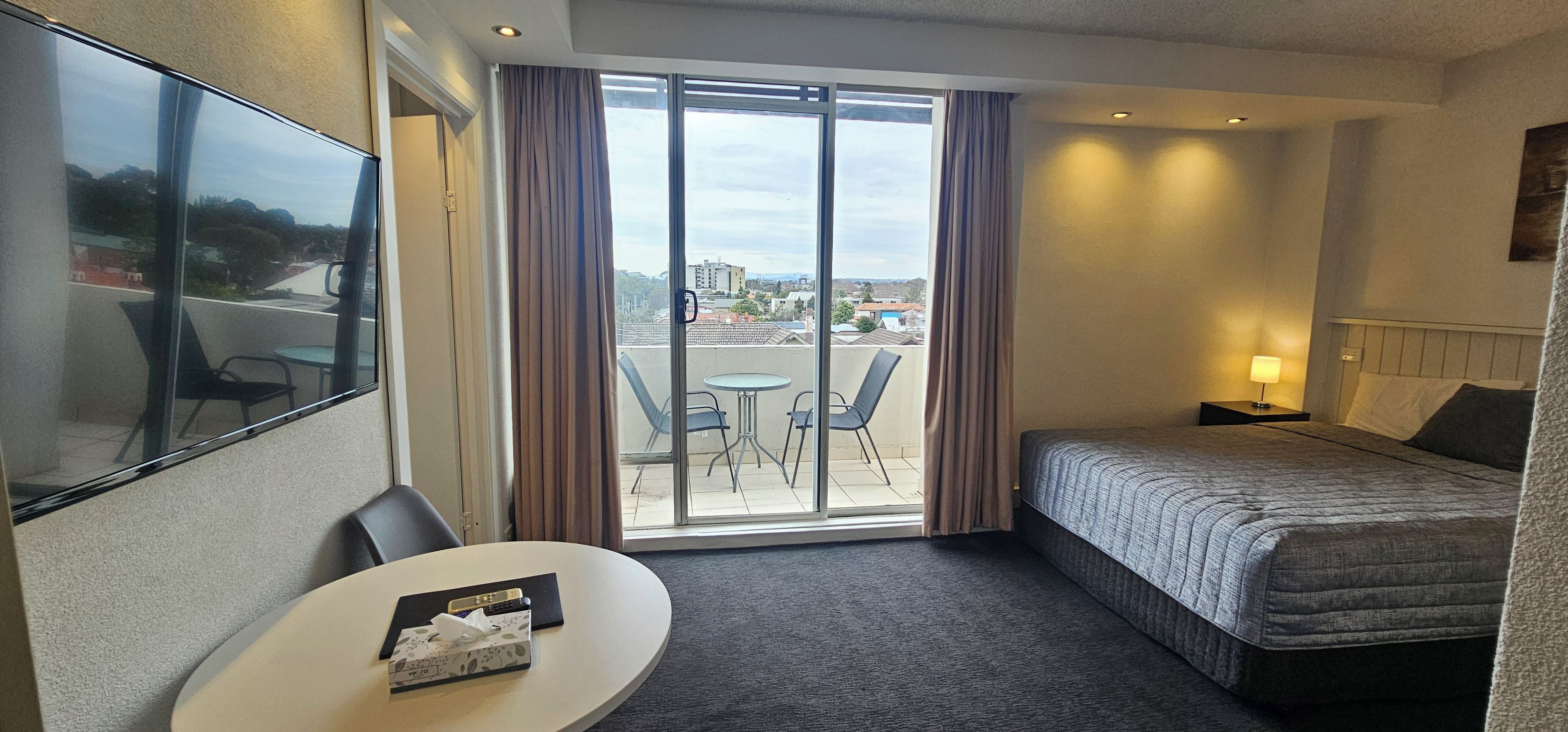 Habitación de hotel con cama, una mesita pequeña con una caja de pañuelos, una silla, una mesita de noche con lámpara y vista a la ciudad a través de un ventanal grande.