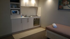 Une cuisine compacte avec des meubles blancs, un four à micro-ondes, une cuisinière et un évier. Un tableau est accroché au mur, et une serviette enroulée est posée sur un banc.