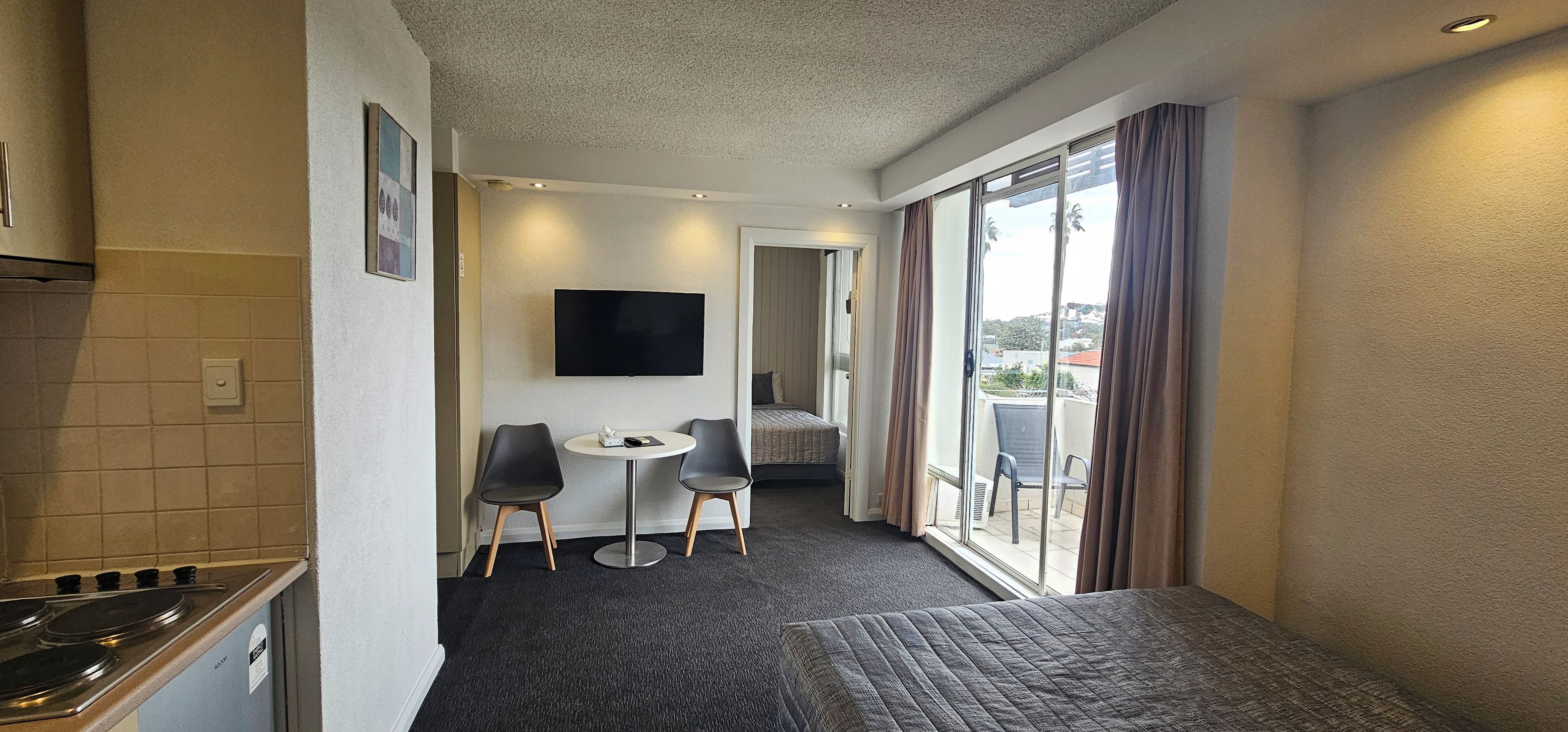 A hotel room with a kitchenette, a flat-screen TV, a small table with two chairs, a bed, and a balcony with curtains.