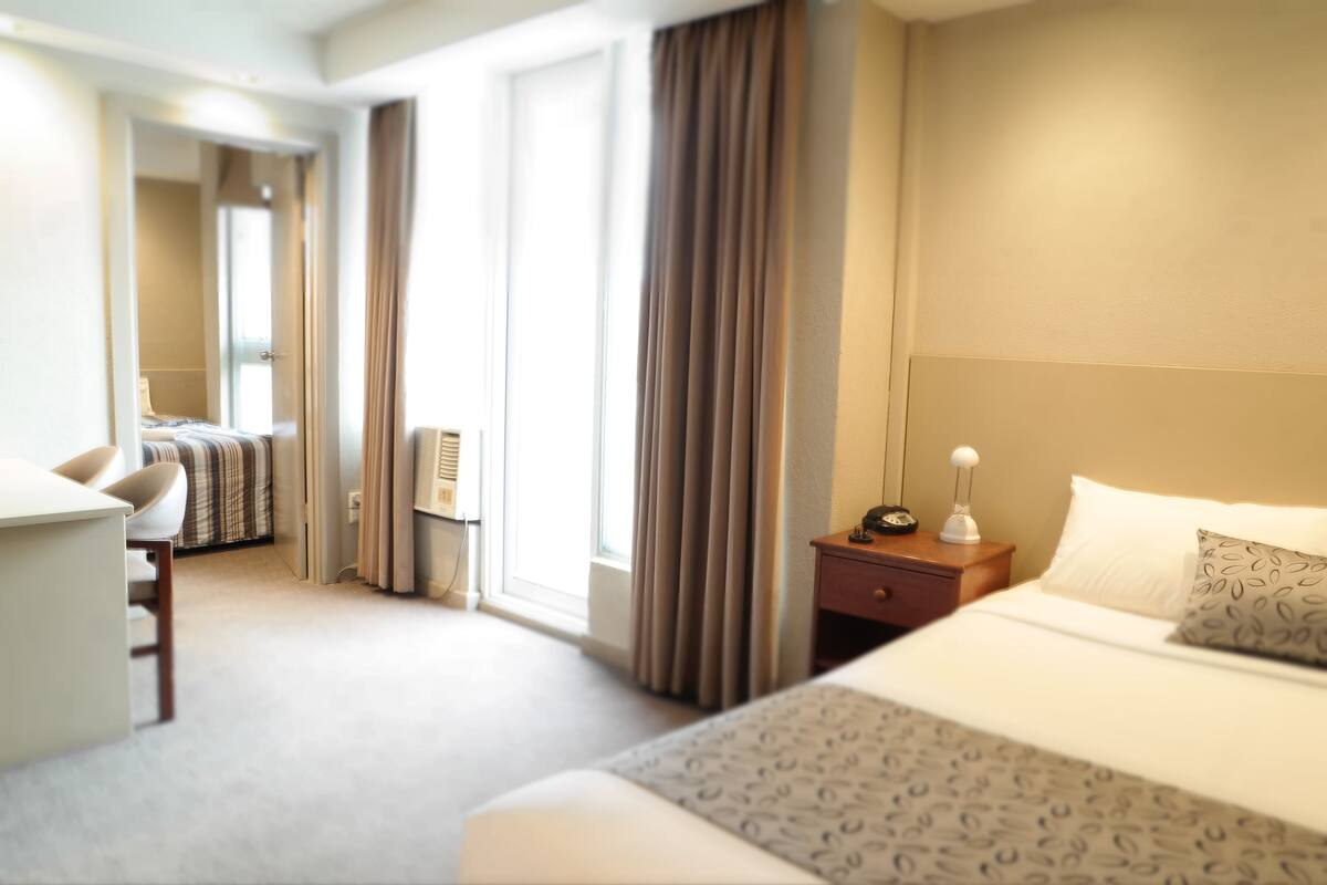 Hotelkamer met een bed, bureau en stoel. Er is een raam met gordijnen, een nachtkastje met een lamp en een wandairconditioner.
