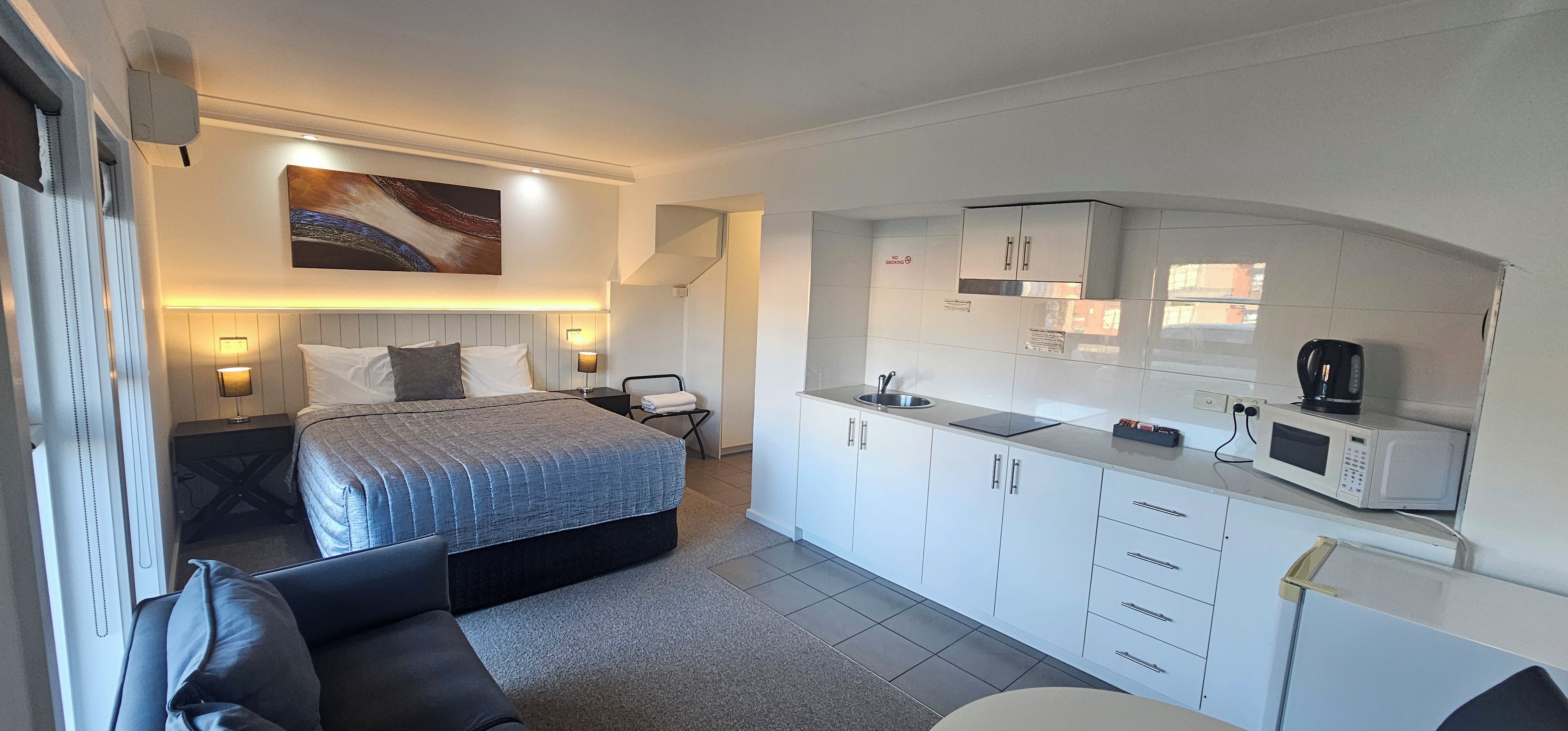 Een kleine hotelkamer met een bed, nachtkastje, lamp en een kitchenette met een magnetron en koffiezetapparaat.