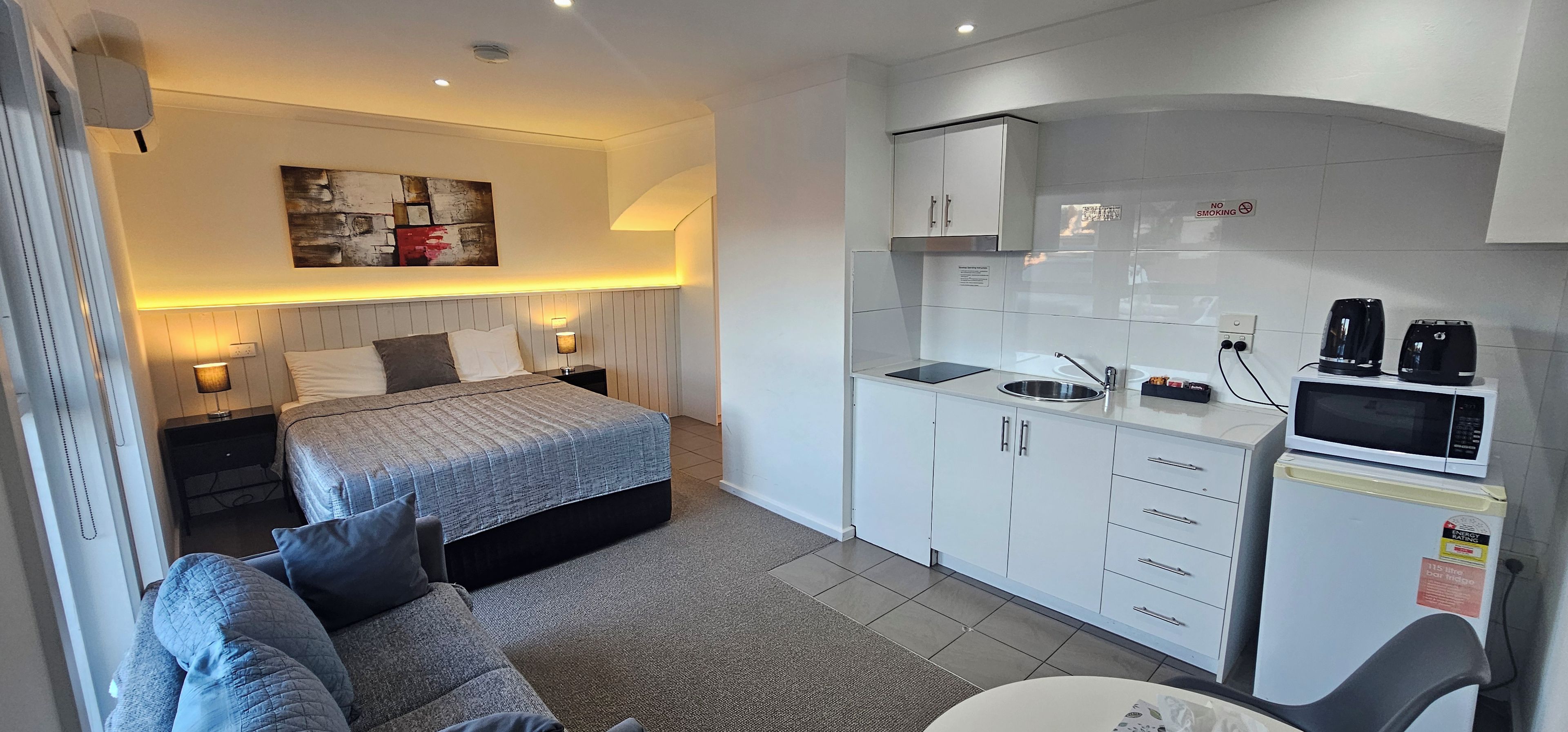 A compact hotel room with a bed, a sofa, a kitchenette, and a small dining area.