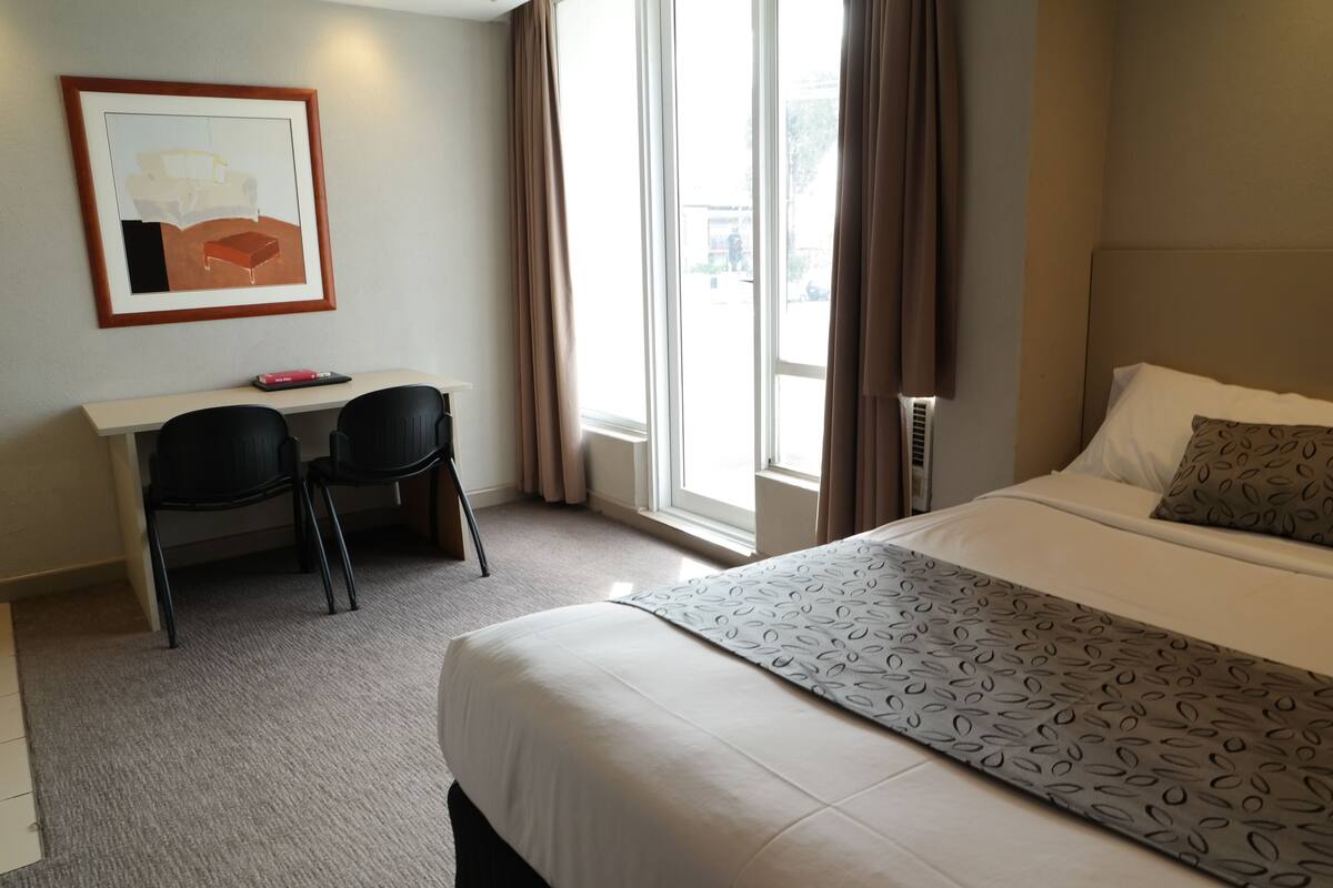 Hotelkamer met een bed, een bureau met twee stoelen, een gekaderd kunstwerk aan de muur en een raam met gordijnen.