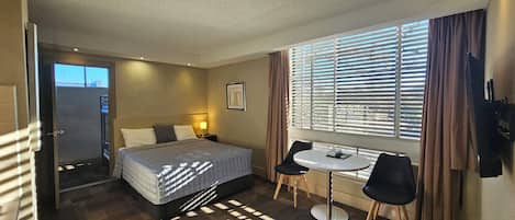 Habitación de hotel con cama, mesita de noche, lámpara, ventana con persianas y una pequeña mesa con dos sillas.