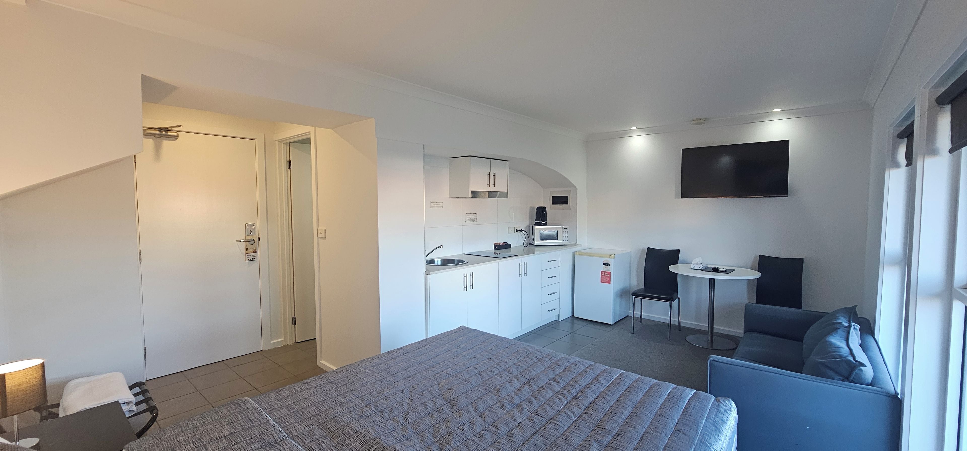 Et moderne hotelværelse med seng, lille tekøkken, siddeområde med bord og stole samt et fjernsyn på væggen.