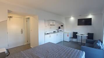 Et moderne hotelværelse med seng, lille tekøkken, siddeområde med bord og stole samt et fjernsyn på væggen.