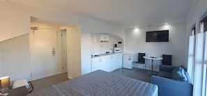 Une chambre d’hôtel moderne équipée d’un lit, d’une petite kitchenette, d’un coin salon avec une table et des chaises, et d’un téléviseur fixé au mur.