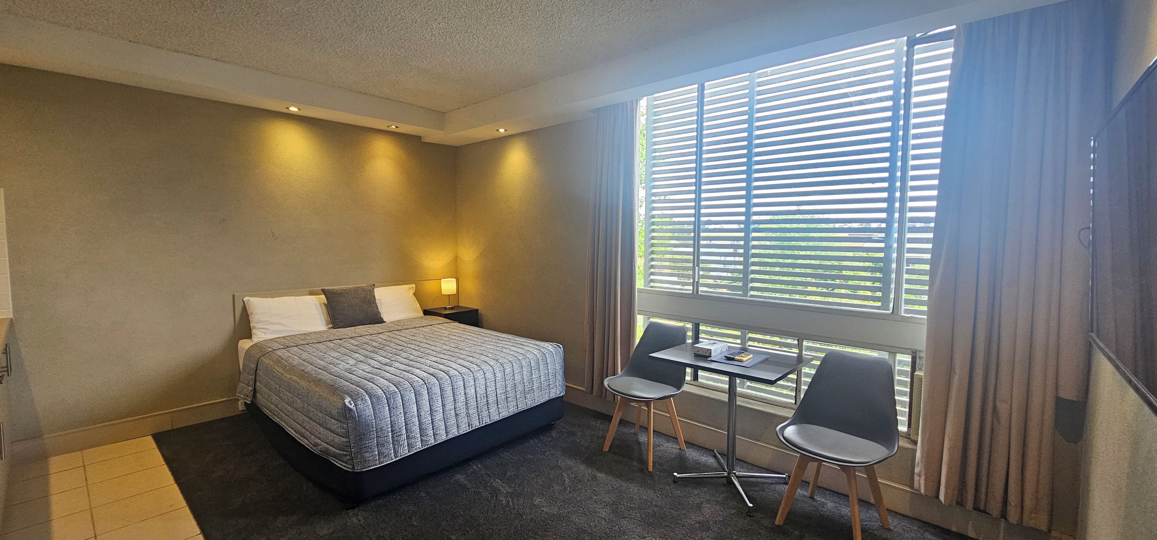 Une chambre d’hôtel comprenant un lit, des tables de chevet, un bureau avec des chaises, une fenêtre avec des stores et un sol recouvert de moquette.