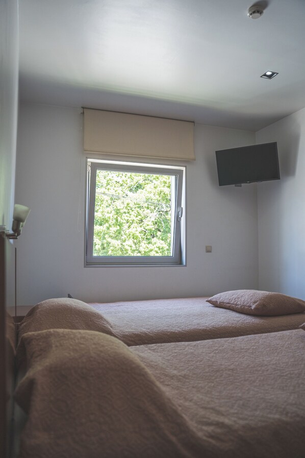 A bedroom with two beds, a window with a view of greenery, a mounted TV, and a ceiling light.