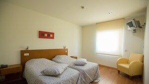 A hotel room with two beds, a television mounted on the wall, a window with blinds, and a chair.