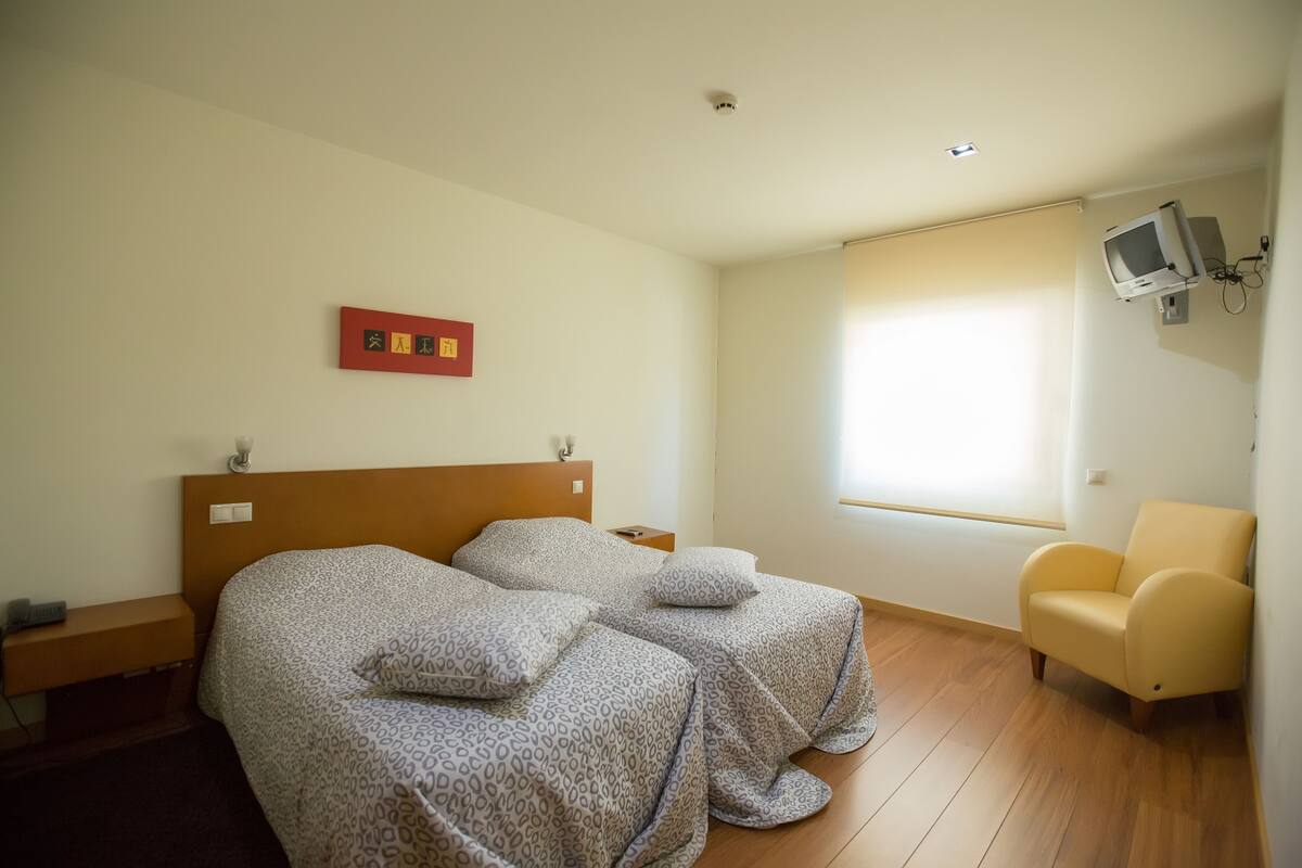 A hotel room with two beds, a television mounted on the wall, a window with blinds, and a chair.