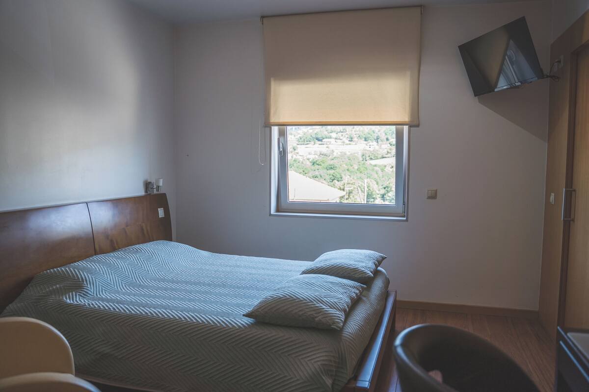 A bedroom with a bed, a window with a view, a TV mounted on the wall, and a wooden cabinet.