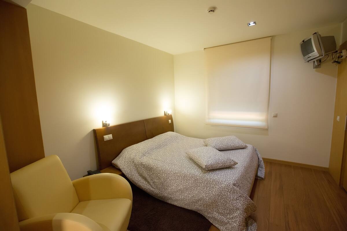 A bedroom with a bed, a chair, a TV mounted on the wall, and a window with blinds.