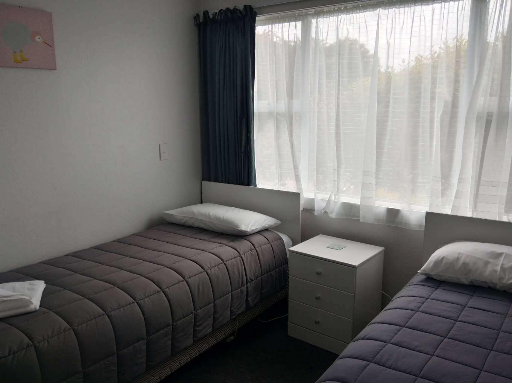 A small, simple room with two beds, a nightstand, and a window with curtains.