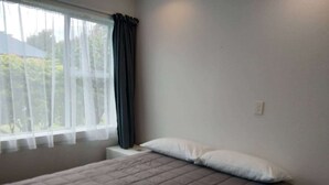 Un dormitorio con cama, ventana con cortinas translúcidas y mesita de noche.