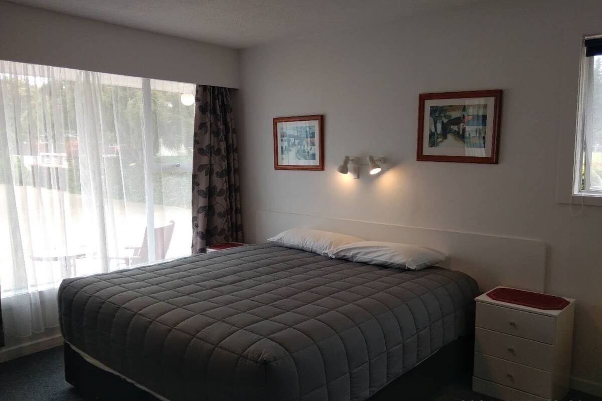 A bedroom with a large bed, a bedside table, a window with curtains, and two framed pictures on the wall.