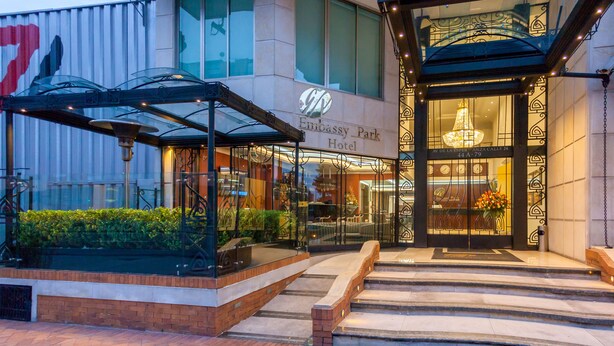 Embassy Park Hotel set udenfor, med glasklædt indgang, et overdækket udendørs siddeområde og en brobelagt sti, der fører til indgangen.