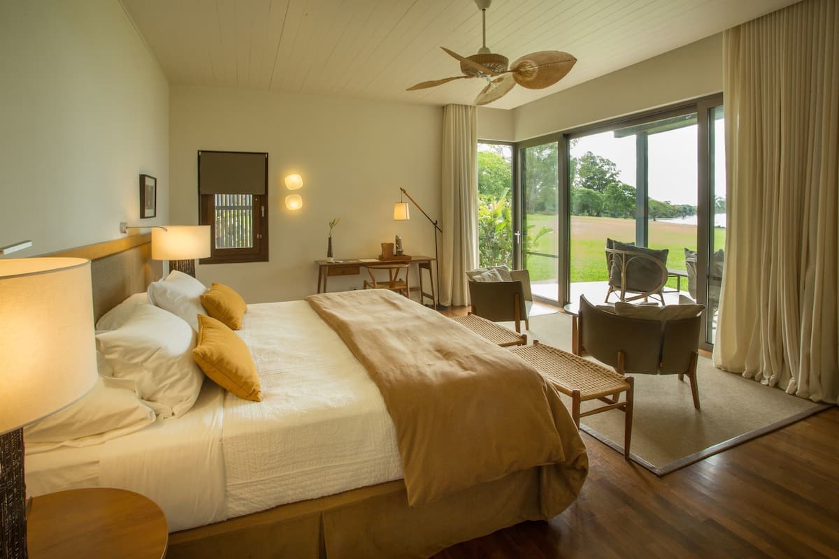 A spacious bedroom with a large bed, a desk, and a chair. There is a view of a garden through the window.