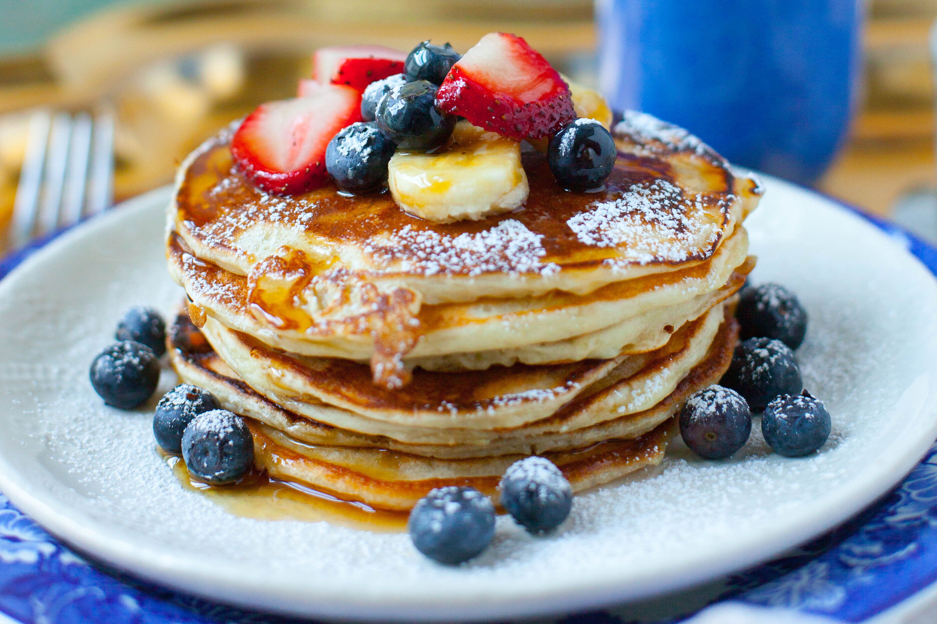 A stack of pancakes with blueberries, strawberries, and a banana on top, served on a plate with syrup and powdered sugar.