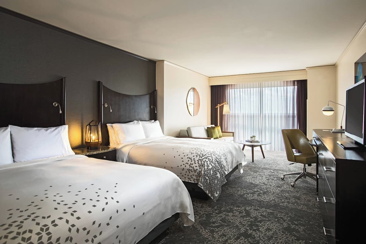 Une chambre d’hôtel avec deux lits, un bureau, une chaise, une télévision, une lampe et une petite table avec une plante.
