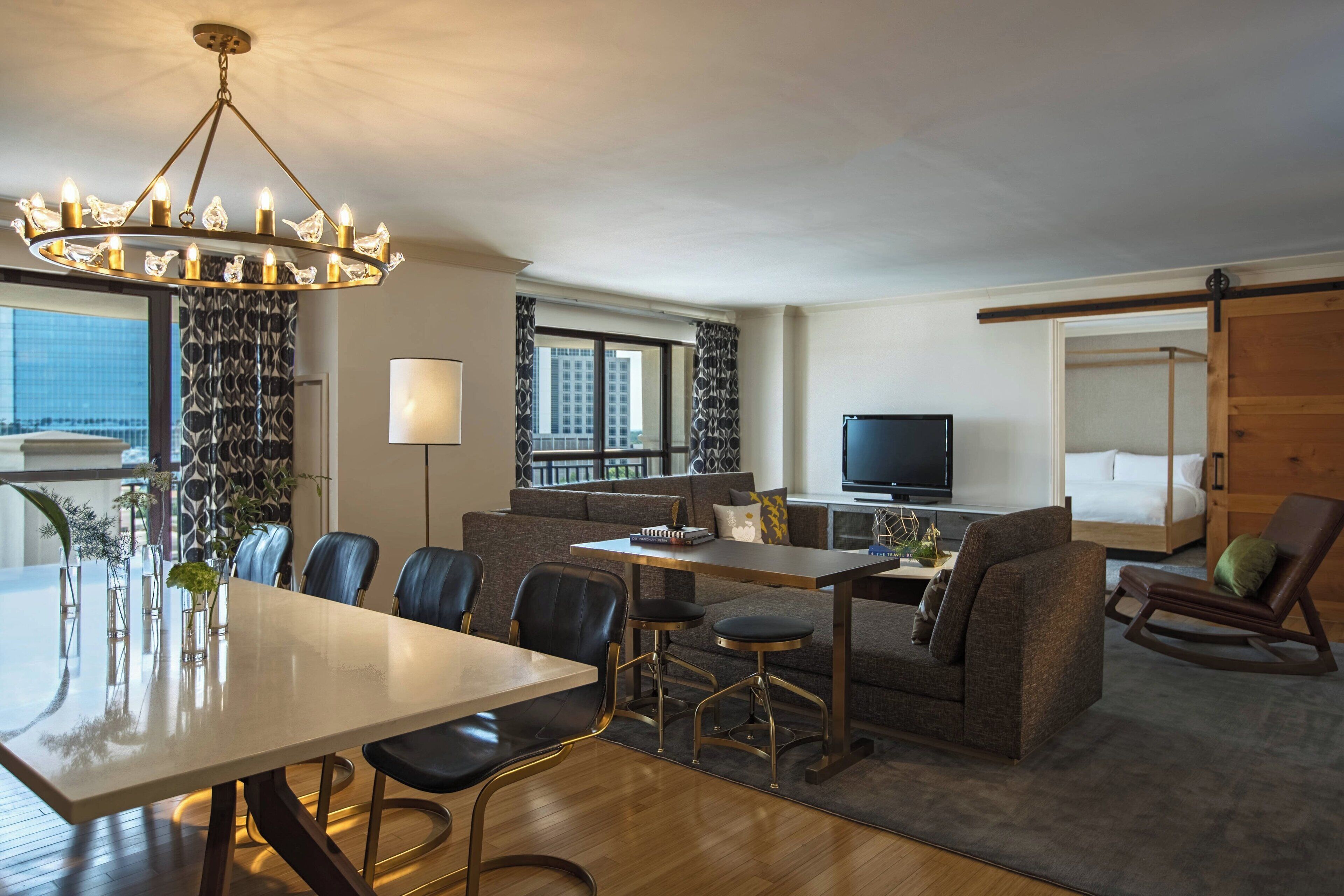 A modern hotel room with a dining area, a sofa, a TV, and a balcony with a city view.