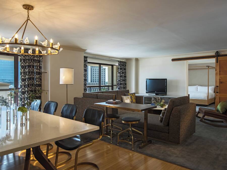 A modern hotel room with a dining area, a sofa, a TV, and a balcony with a city view.