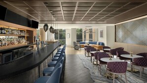A modern bar area with a long counter, bar stools, and a variety of liquor bottles. There are several seating areas with tables and chairs, and a large window providing a view of the outdoors. - Renaissance Raleigh North Hills Hotel (Raleigh)