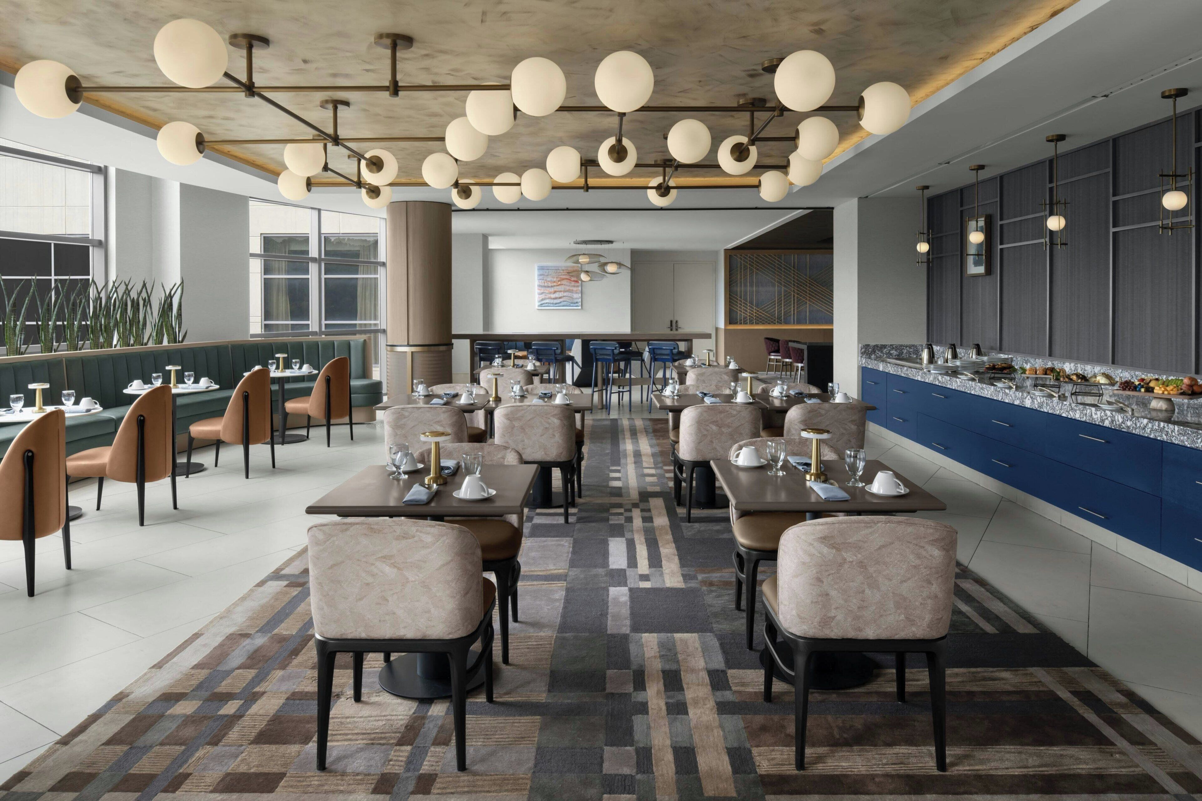 Un restaurante moderno con una barra larga, varias mesas y sillas. Hay luces colgantes y una alfombra estampada.