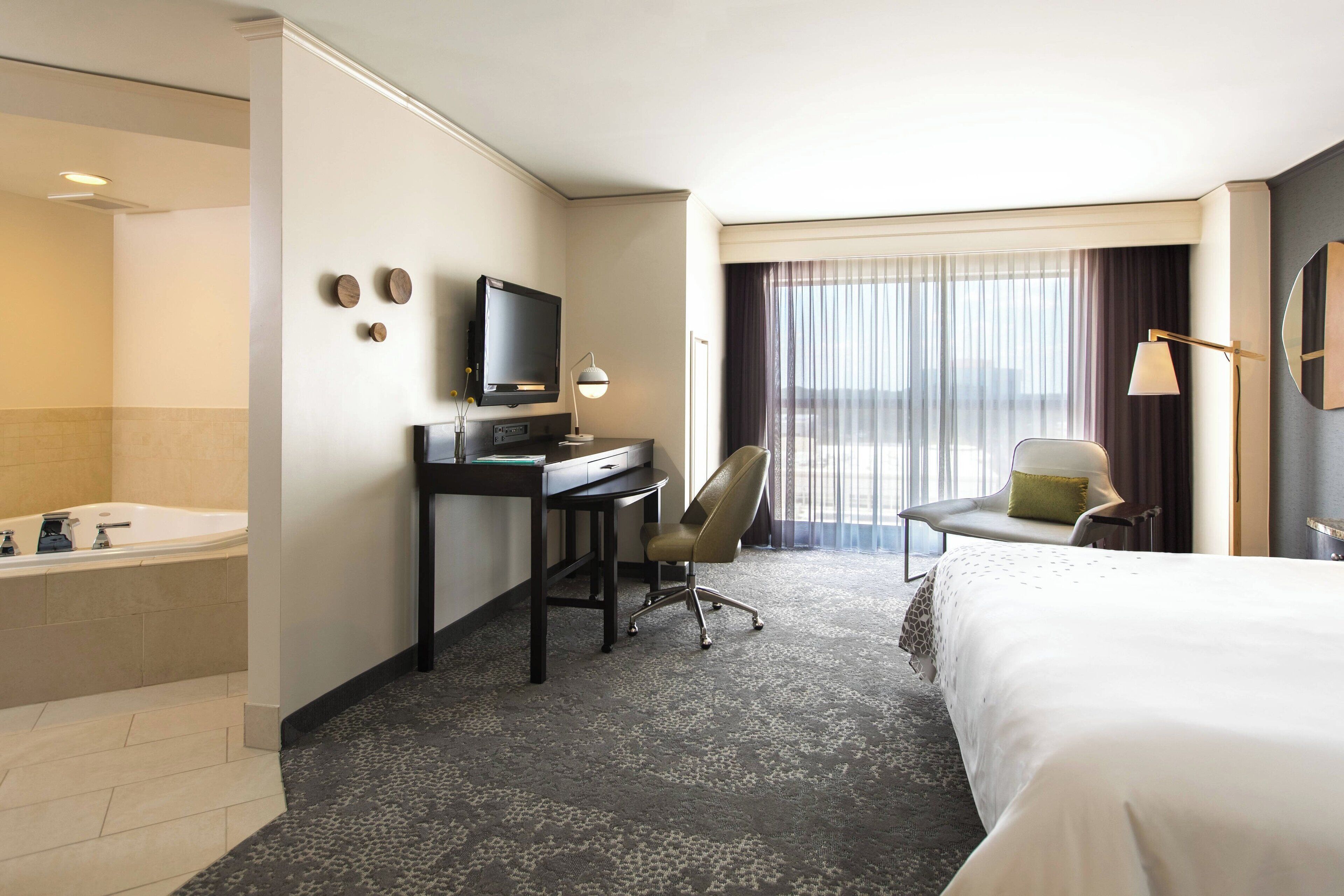 Habitación de hotel con cama, escritorio con computadora, silla, televisor y un amplio ventanal con cortinas.