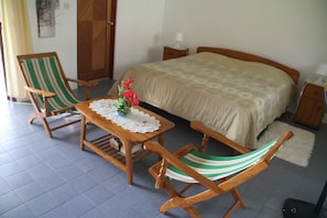 Quarto com cama, duas cadeiras de madeira, uma mesa pequena com toalha de mesa florida e um criado-mudo.