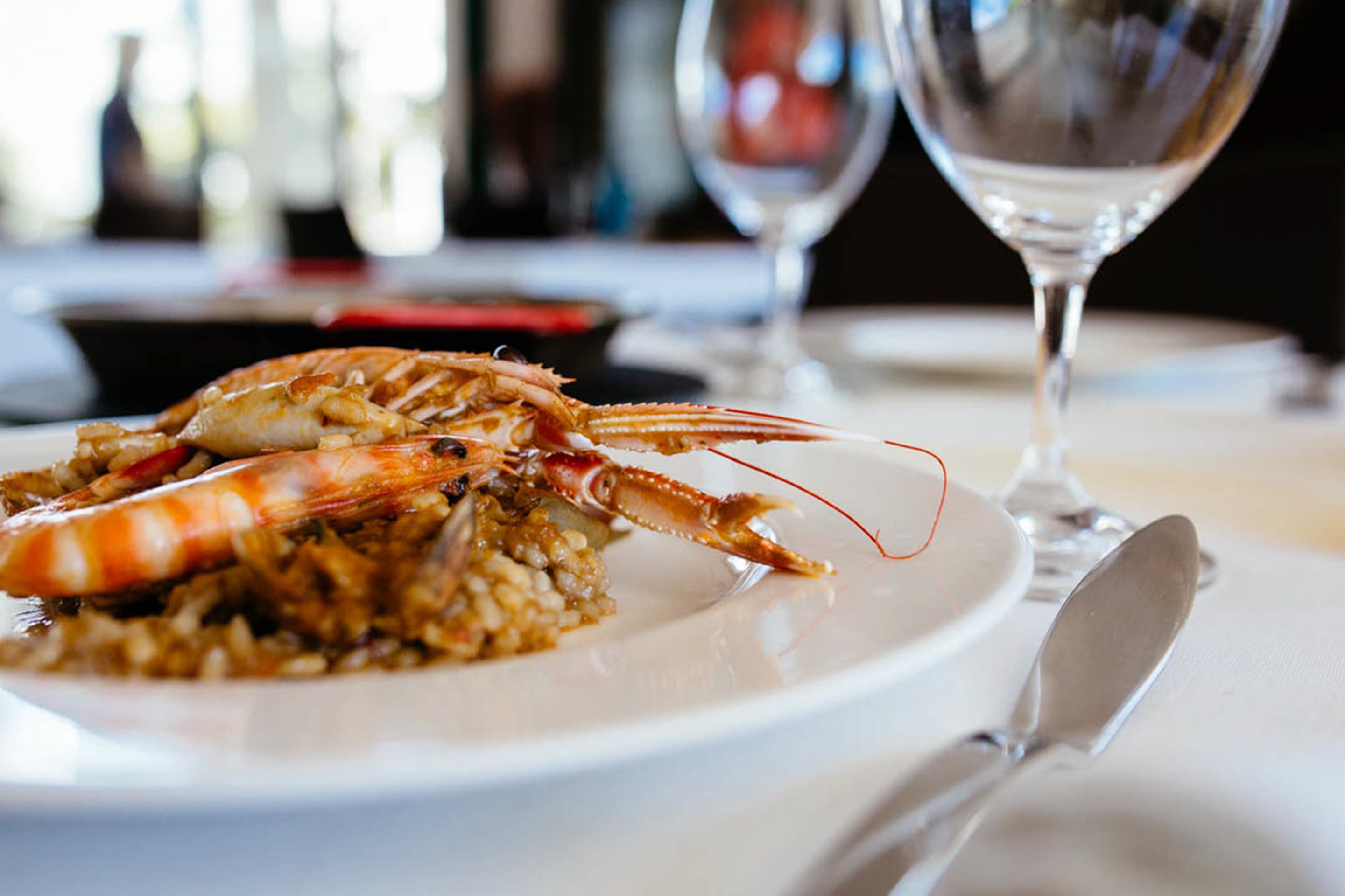 Un plato de comida con un langostino, arroz, una copa de vino y un tenedor sobre una mesa.