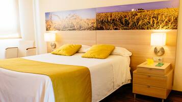 Quarto de hotel com uma cama, duas luminárias de cabeceira, uma mesa de cabeceira com luminária e uma obra de arte na parede retratando campos de trigo.