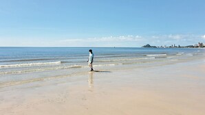 A beach with a person walking along the shore. The beach is wide and sandy with gentle waves. In the background, there is a distant cityscape with buildings and a small hill. - Thipurai city hotel (Hua Hin)