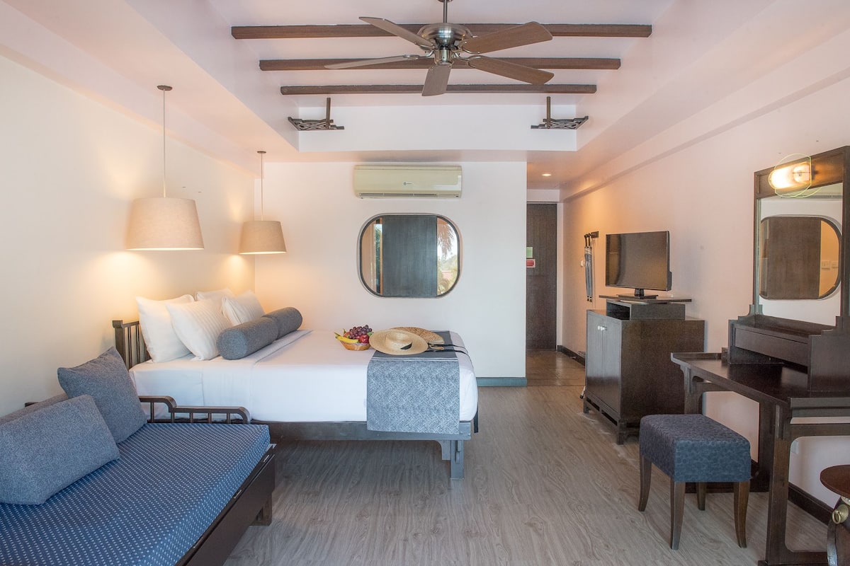 A hotel room with a bed, a desk, a chair, a ceiling fan, and a TV.