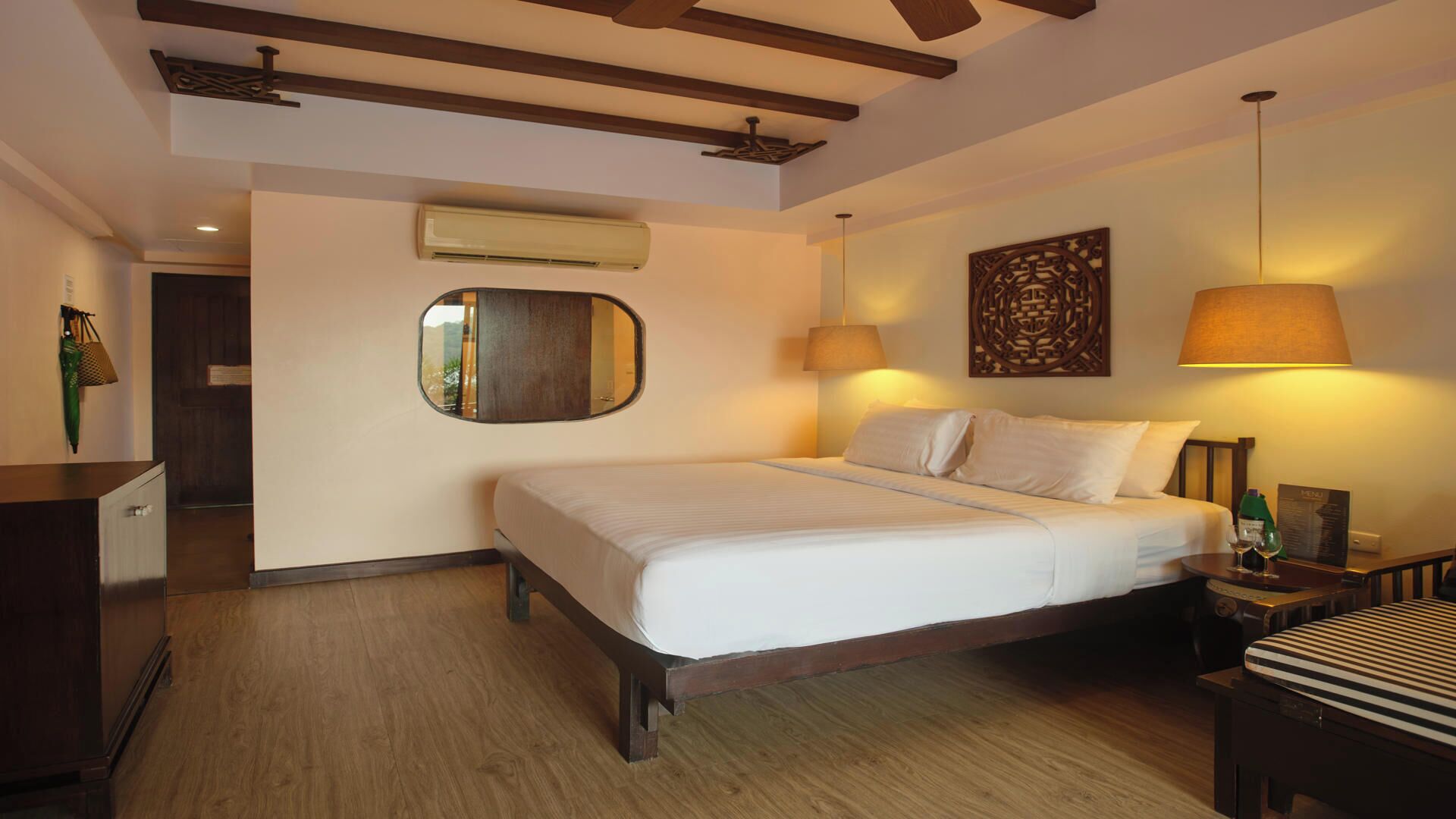 A hotel room with a bed, bedside lamps, a mirror, an air conditioning unit, and a wooden ceiling.