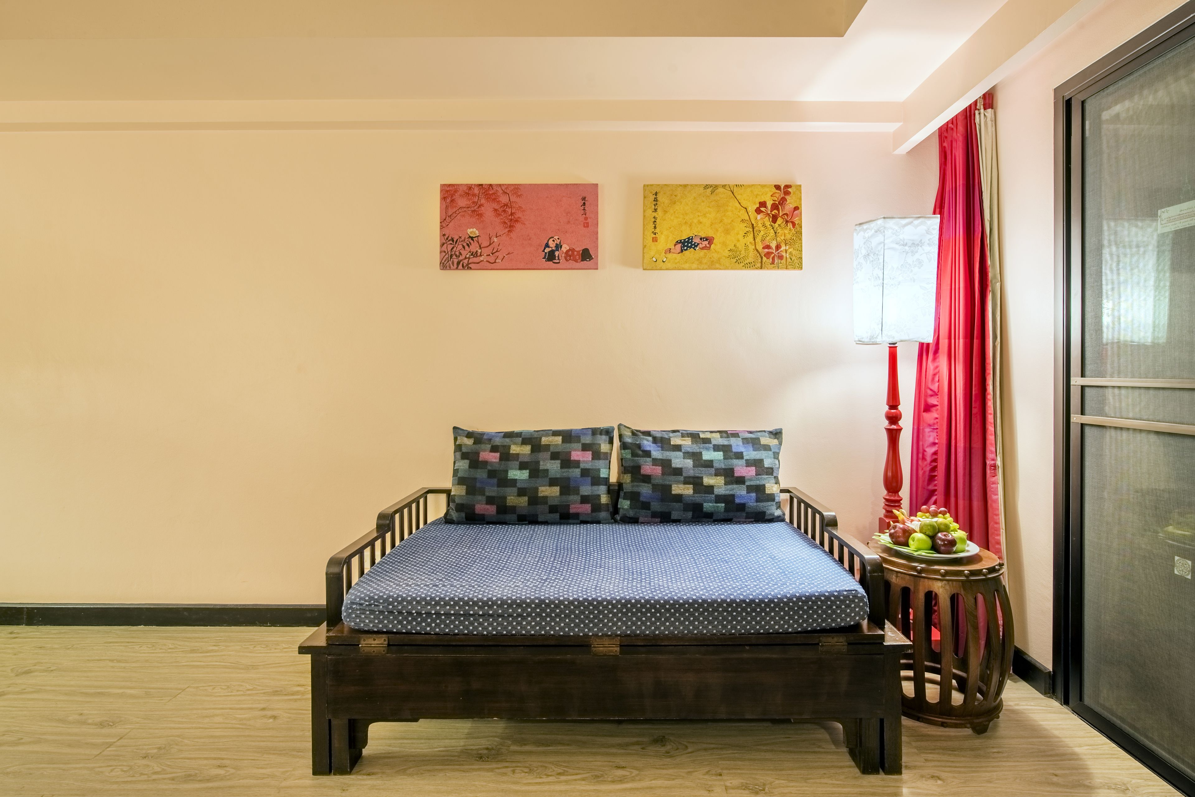 A bed with a blue patterned bedspread and two pillows, a small wooden side table with fruit, and two wall-mounted artworks.