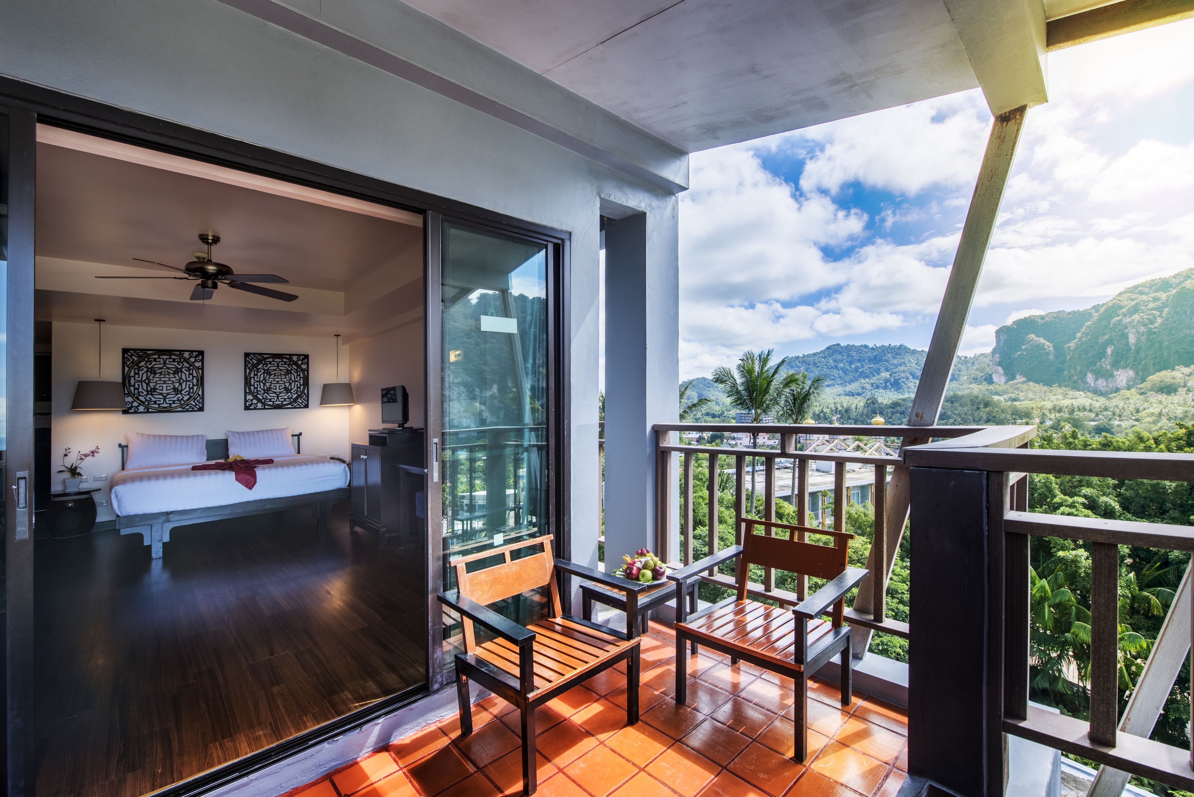 A balcony with a view of a mountainous landscape, a bedroom with a bed and ceiling fan, and a dining area with chairs and a table.