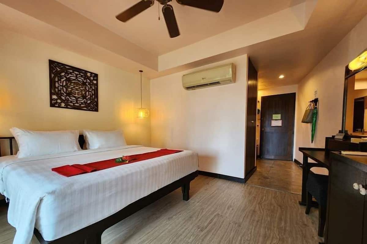 A hotel room with a bed, a ceiling fan, an air conditioning unit, a desk with a mirror, and a wall-mounted artwork.