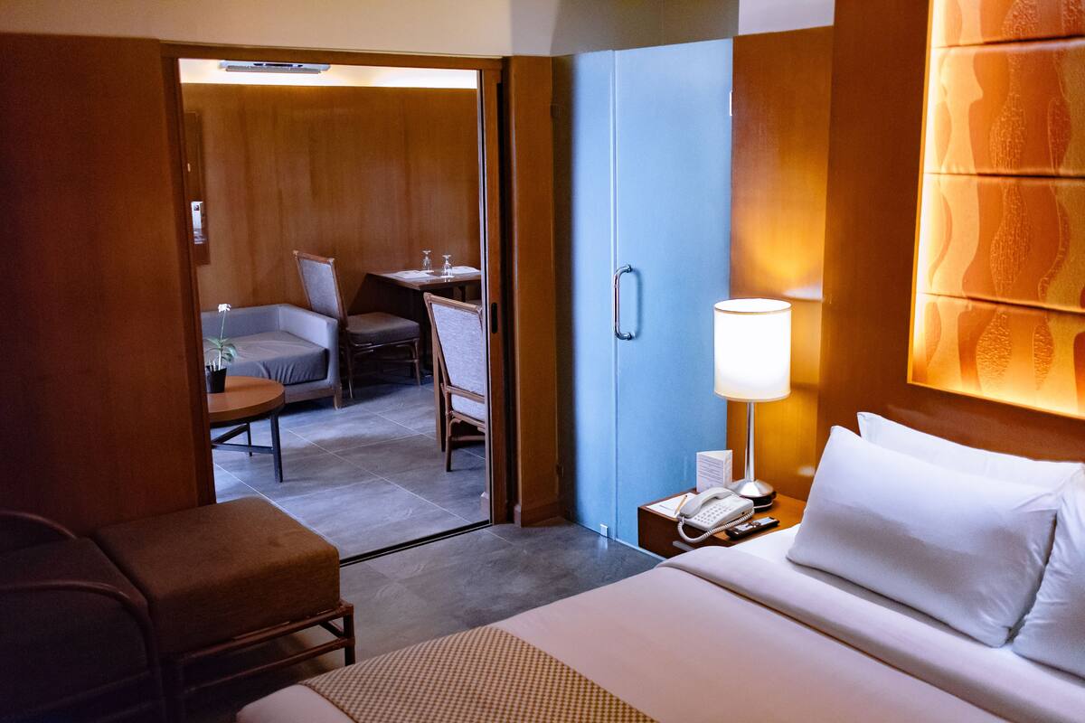 A hotel room with a bed, bedside table with a lamp, a chair, a desk with a chair, and a door leading to a dining area.