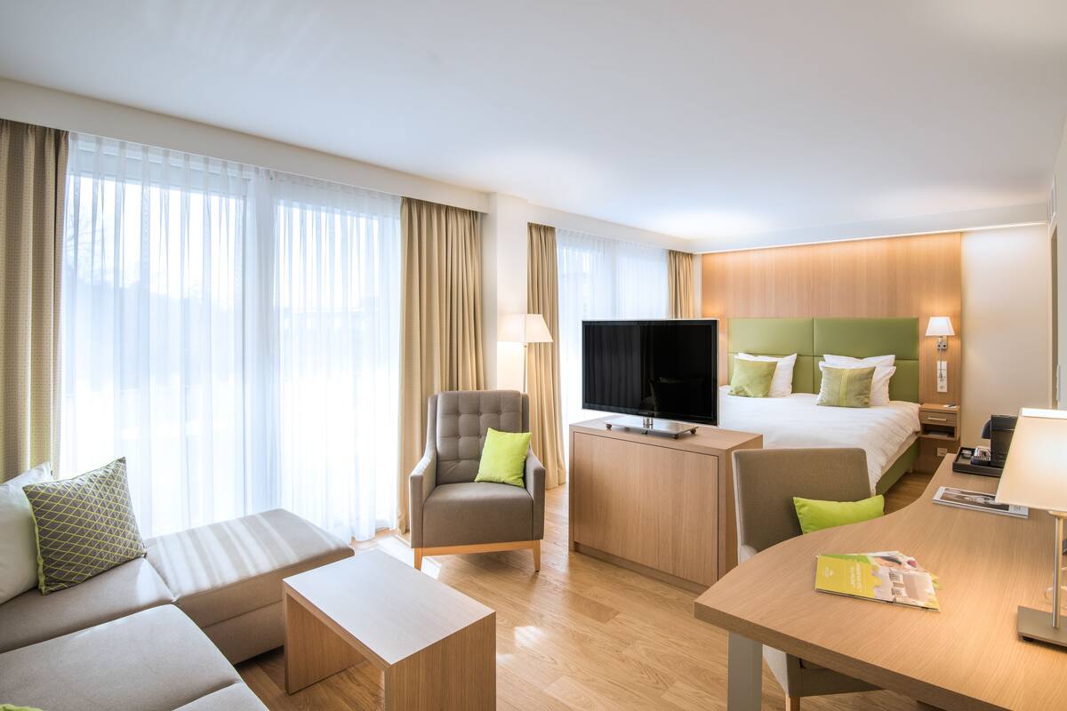 Habitación de hotel con sofá, sillón, televisión, cama y escritorio con lámpara.