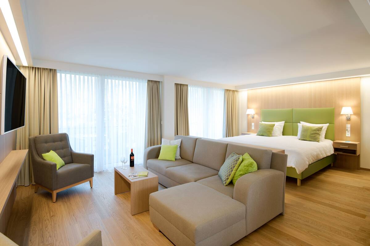 Una habitación de hotel moderna con sofá, sillón y cama con cabecero verde.