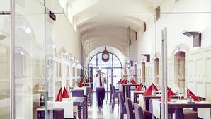 A spacious restaurant with high ceilings and a central arched passageway. Tables are set with red napkins and wine glasses.