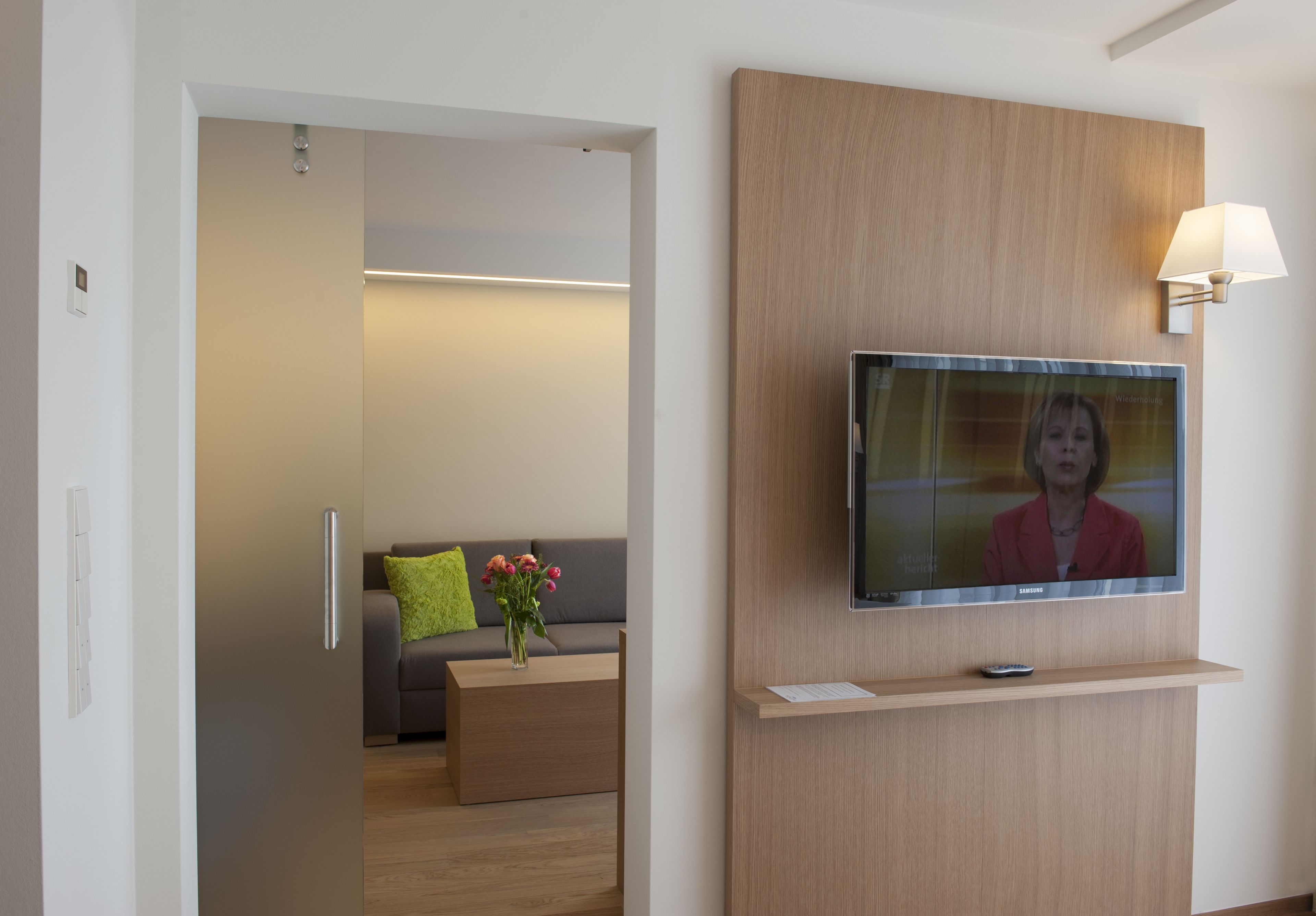 A modern hotel room with a flat-screen TV mounted on the wall, a wooden console table, a sofa with a green pillow, and a vase with flowers.