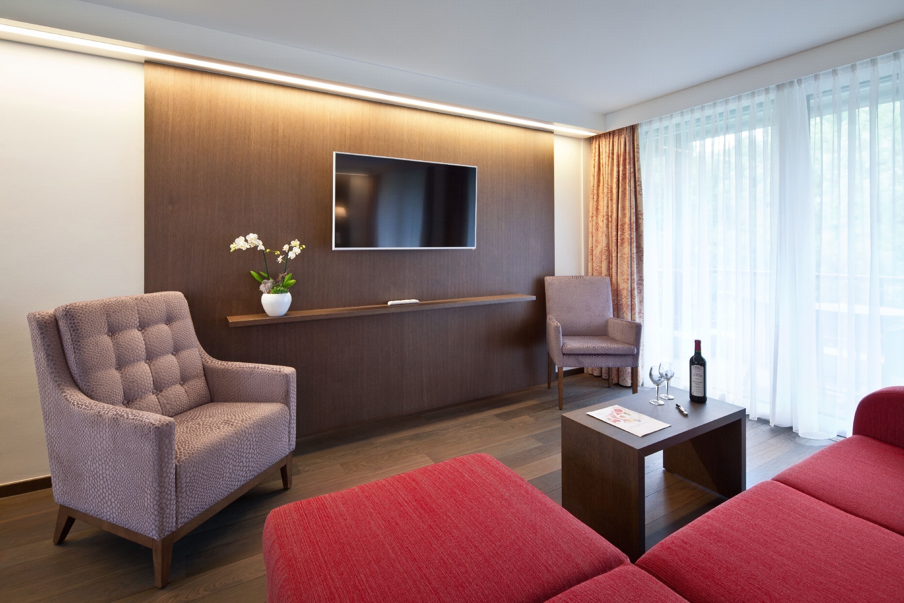 A modern living room with a flat-screen TV mounted on a wooden wall, two armchairs, a red sofa, a coffee table, and a potted plant.